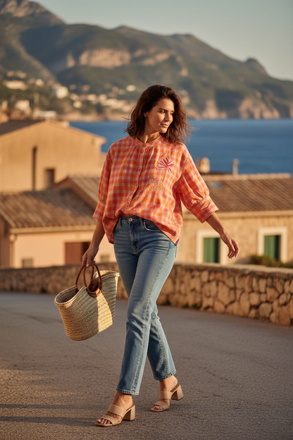 Fashion editorial image of a woman with a natural, effortless Mediterranean aesthetic, inspired by 70s French Riviera style. She has soft, slightly messy hair moved gently by the sea breeze, natural glowing skin, minimal makeup, and a subtle, spontaneous smile.

She is wearing a relaxed, boxy shirt in lightweight cotton with small orange and nude check pattern, featuring elbow-length subtly puffed sleeves with soft volume and movement.

The shirt is styled with high-waisted vintage blue denim jeans (effortl